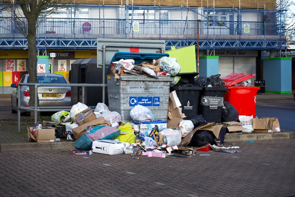 A large collection of mixed household rubbish and waste materials is piled on a paved street near a small parking area, with several overflowing black and red plastic bins, a grey recycling container marked for paper and card, and various cardboard boxes and paper parcels strewn across the ground. The waste includes crumpled paper, flattened cardboard boxes, plastic bags, and packaging, with some items partially out of the bins or slightly leaning over. In the background, there are commercial storefronts with window displays, signage, and a blue metal scaffolding structure covering the upper part of a building. A mature tree is situated on the left, and a metal railing separates the waste area from parked vehicles, including a silver car. The scene is illuminated by natural daylight, creating a neutral, everyday view of street-side rubbish accumulation, illustrating the type of waste collection handled by private waste management services like Rubbish Collection Chelsea, which provides alternative rubbish removal options outside standard council collection.