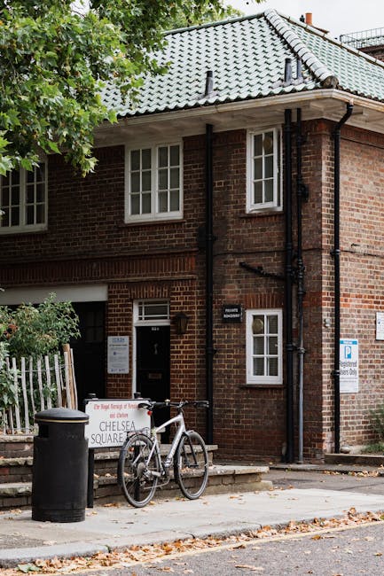 The image depicts the exterior of a two-storey brick building with a pitched gray tiled roof, situated on a residential street. In the foreground, there is a concrete sidewalk with scattered fallen leaves and a black cylindrical trash bin positioned near the curb. Leaning against the building's front wall, slightly to the left, is a white bicycle with black handlebars and seat, locked to a metal stand. Adjacent to the bicycle, there's a signboard indicating a public parking zone, partially obscured by green foliage from a nearby tree with dense leaves extending into the upper left corner of the frame. The building features white-framed sash windows, with two larger windows on the upper floor and a smaller window near the door, to the right of the entrance. The door, located in the center, is dark-colored and has a mailbox and a doorbell next to it. To the right of the entrance, there is a small, rectangular sign attached to the brick wall, and beside it, a larger parking sign indicating permit requirements, mounted on a metal post. The scene is lit by natural daylight, with soft shadows cast on the brickwork and pavement, representing a typical urban setting where private waste collection services like Rubbish Collection Chelsea facilitate alternative waste handling for residents. The overall environment appears clean and orderly, with no visible rubbish or debris in the vicinity.