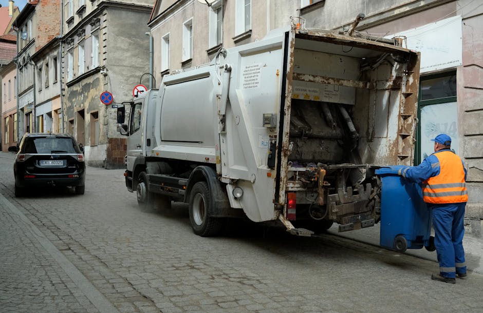 A large collection of mixed household rubbish and waste materials is piled on a paved street near a small parking area, with several overflowing black and red plastic bins, a grey recycling container marked for paper and card, and various cardboard boxes and paper parcels strewn across the ground. The waste includes crumpled paper, flattened cardboard boxes, plastic bags, and packaging, with some items partially out of the bins or slightly leaning over. In the background, there are commercial storefronts with window displays, signage, and a blue metal scaffolding structure covering the upper part of a building. A mature tree is situated on the left, and a metal railing separates the waste area from parked vehicles, including a silver car. The scene is illuminated by natural daylight, creating a neutral, everyday view of street-side rubbish accumulation, illustrating the type of waste collection handled by private waste management services like Rubbish Collection Chelsea, which provides alternative rubbish removal options outside standard council collection.