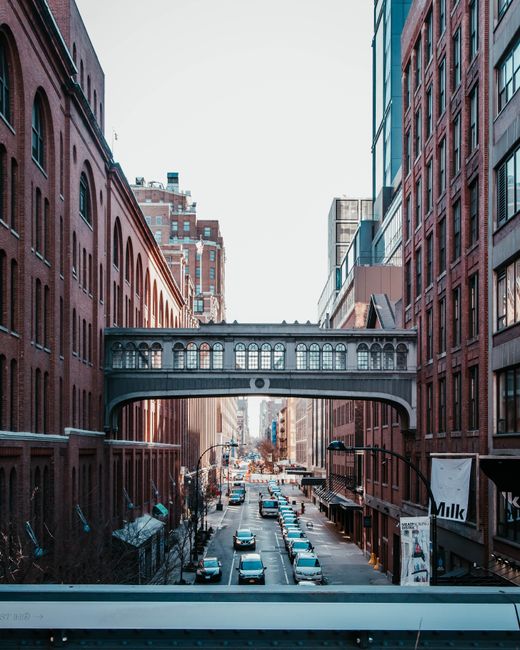 A city street viewed from an elevated position, with a narrow road lined by historic red-brick buildings on both sides. An ornate, dark metal bridge spans across the street, connecting the buildings on each side, featuring decorative arches and windowed panels. The street below is busy with parked cars and moving vehicles, extending into the distance with additional buildings and streetlights visible under a clear, bright sky. The surrounding environment suggests an urban area with a mix of old and modern architecture, emphasizing the contrast between traditional brick facades and contemporary glass elements. The scene exhibits natural daylight, highlighting the textures of brickwork and asphalt, and reflecting off vehicle surfaces. In the context of waste management and rubbish removal services, the image illustrates a typical cityscape where private rubbish collection or on-site clearance could be relevant, especially for commercial or residential property maintenance. The presence of parked cars and urban infrastructure underlines the importance of efficient rubbish handling in busy city environments, aligning subtly with alternative waste management options within modern urban areas.