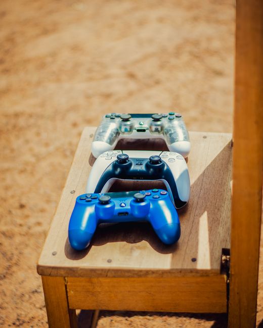 Three video game controllers are positioned on a light brown wooden table, displaying a top-down view in a well-lit indoor environment. The front controller is bright blue with a textured grip and black buttons, while the middle controller is dark blue with a matte finish and black buttons. The third controller at the back is semi-transparent with a clear casing revealing internal components, and features a similar button layout. The table is situated in a space with beige carpet flooring and a plain, textured beige wall in the background. Shadows cast by the controllers suggest natural or ambient lighting. This arrangement of gaming controllers may be part of a recreational area intended for leisure activities, and while not directly related to rubbish removal services, such settings could be encountered in environments requiring on-site waste clearance or cleaning, often provided by independent waste management providers like Rubbish Collection Chelsea, to maintain tidy and functional living or gaming spaces.