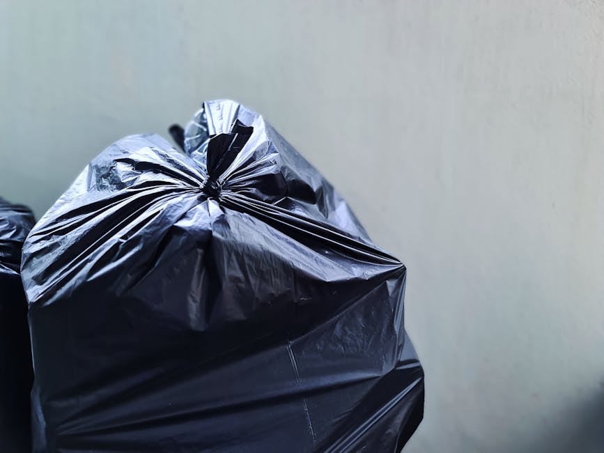 Close-up of several tightly tied black rubbish bags made from glossy plastic, placed against a plain, light-colored wall. The bags appear to be filled with waste, with some showing creases and folds on their surface, and are positioned on a surface, possibly a floor or a collection point, with minimal surrounding context visible. The lighting highlights the reflective quality of the plastic material, emphasizing the texture and contours of each bag. The scene represents typical waste ready for collection by a private rubbish removal service such as Rubbish Collection Chelsea, illustrating an instance of on-site waste disposal that may be part of ongoing rubbish management or clearance activities.