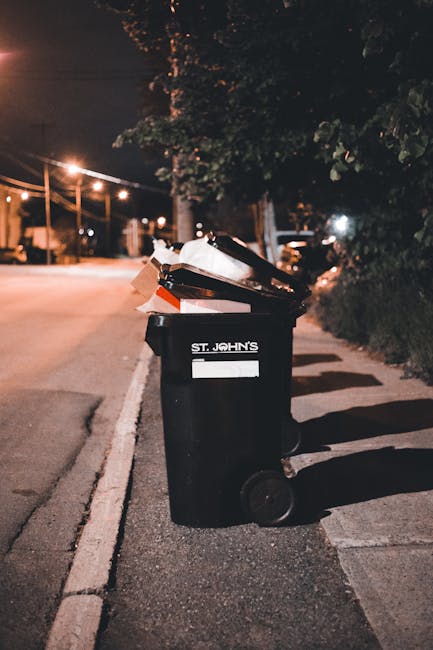A black wheeled rubbish bin with a white label and the words 'ST. JOHN'S' printed on it is positioned on a sidewalk next to the road in an urban area during nighttime. The bin's lid is open, revealing various waste items, including cardboard boxes and plastic containers, some of which are protruding from the top. Streetlights cast a warm glow on the scene, illuminating the surrounding pavement, nearby trees, and bushes. In the background, there are faint outlines of parked cars and buildings, with additional streetlights stretching along the road, creating a softly lit environment suitable for independent waste collection or private rubbish removal services. The overall setting suggests a typical street scene where waste has been temporarily placed outside for collection, aligned with the context of alternative waste handling solutions like those offered by Rubbish Collection Chelsea.
