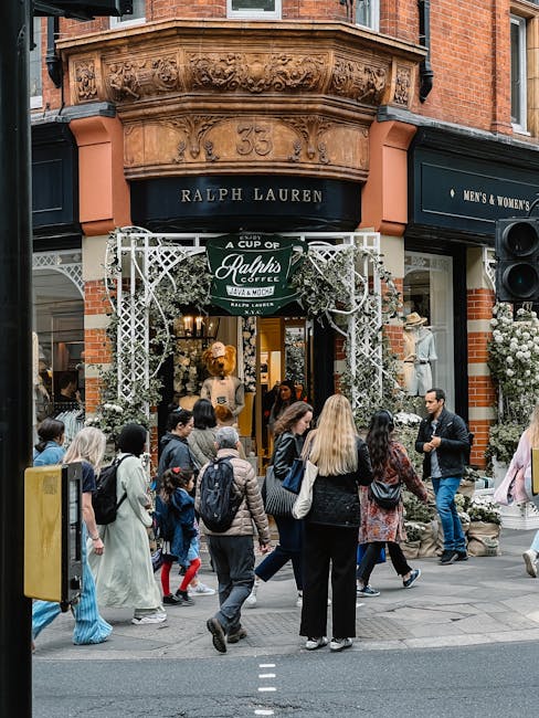 The exterior of a Ralph Lauren store on a busy street corner, featuring a classic brick facade with decorative stone detailing above the entrance displaying the number '55'. The entrance is framed by white decorative metalwork with floral accents and has a dark awning with the Ralph Lauren name. A large, dark sign on the right indicates men's and women's clothing, and the store window displays mannequins dressed in fashionable attire. In front of the store, a diverse group of pedestrians, including men, women, and children, are walking and standing on the sidewalk, some carrying backpacks and wearing casual clothing. The scene is set during daylight, with natural light illuminating the street and store frontage. The area appears lively and well-frequented, reflecting a typical retail environment suitable for independent or private waste collection and rubbish removal services for commercial premises.