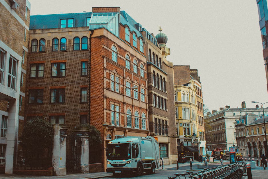 A street scene featuring a mixture of historic and modern buildings with various architectural styles, including brick facades with arched and rectangular windows, and detailed cornices. In the foreground, a white refuse collection truck with a compact design is parked on the pavement, partially blocking the view of a building entrance enclosed by a black wrought iron gate supported by stone pillars. The truck is positioned close to the curb, indicating recent rubbish collection activity. To the right, there is a metal bike rack with several bicycles parked, and the street is lined with bollards separating the pedestrian walkway from the road. The environment is urban, with other vehicles and street furniture visible further down the street, under an overcast sky. This scene exemplifies independent waste collection services operating within a city context, with Rubbish Collection Chelsea providing private rubbish removal for local businesses and households in the area, supporting alternative waste handling outside of municipal systems.