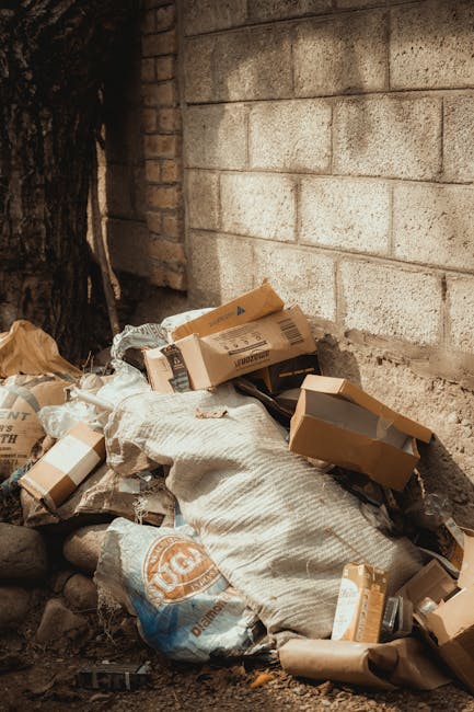 A pile of mixed waste materials, including flattened cardboard boxes, paper bags, and plastic packaging, is situated against a rough concrete brick wall outdoors. The cardboard boxes vary in size and are partially opened or crumpled, with some showing printed labels or barcode markings. The paper bags appear used and crumpled, with visible folds and creases. A large, white woven sack is also present, partially filled with smaller debris. The objects rest on an uneven surface composed of dirt and small stones, with a nearby tree trunk partially visible on the left side of the frame. The scene is illuminated by natural daylight, casting soft shadows, and suggests an outdoor setting typical of a private property or alleyway where waste has been temporarily collected for disposal or independent waste handling by companies such as Rubbish Collection Chelsea. The wall behind the rubbish is unpainted, made of concrete blocks, providing a neutral backdrop that emphasizes the cluttered nature of the waste pile without any additional background context.