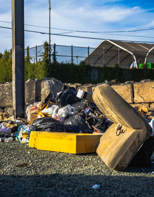 A mound of mixed waste materials is piled on an asphalt surface, with black plastic rubbish bags, a yellow plastic container, and an old, dusty car seat with fabric upholstery. The waste appears to include discarded packaging, paper, and other miscellaneous debris. Behind the pile, there are irregularly cut stone blocks forming a low retaining wall, with a metal pole and a concrete wall in the background. Further behind, a chain-link fence and lush green foliage are visible, with a clear blue sky overhead. To the right, part of a large, curved metal structure, possibly a sports stadium or a covered area, can be seen. The scene is lit by natural daylight, highlighting the textures of the trash, stones, and surrounding environment. The image provides an example of an outdoor rubbish accumulation, illustrating the type of waste that services like Rubbish Collection Chelsea may handle through independent collection or on-site clearance, contrasting with council waste disposal.
