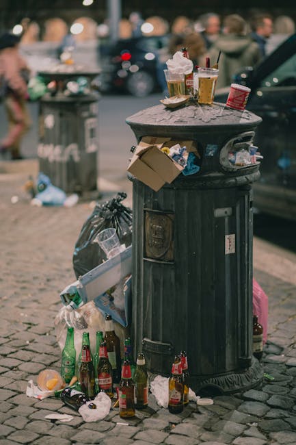A pile of mixed waste materials, including flattened cardboard boxes, paper bags, and plastic packaging, is situated against a rough concrete brick wall outdoors. The cardboard boxes vary in size and are partially opened or crumpled, with some showing printed labels or barcode markings. The paper bags appear used and crumpled, with visible folds and creases. A large, white woven sack is also present, partially filled with smaller debris. The objects rest on an uneven surface composed of dirt and small stones, with a nearby tree trunk partially visible on the left side of the frame. The scene is illuminated by natural daylight, casting soft shadows, and suggests an outdoor setting typical of a private property or alleyway where waste has been temporarily collected for disposal or independent waste handling by companies such as Rubbish Collection Chelsea. The wall behind the rubbish is unpainted, made of concrete blocks, providing a neutral backdrop that emphasizes the cluttered nature of the waste pile without any additional background context.
